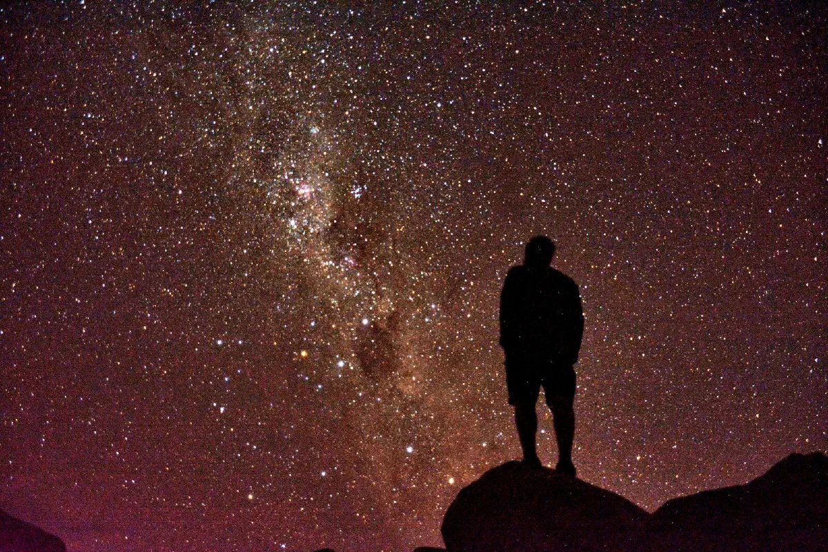 A lone figure gazing up at the Milky Way over a dark landscape